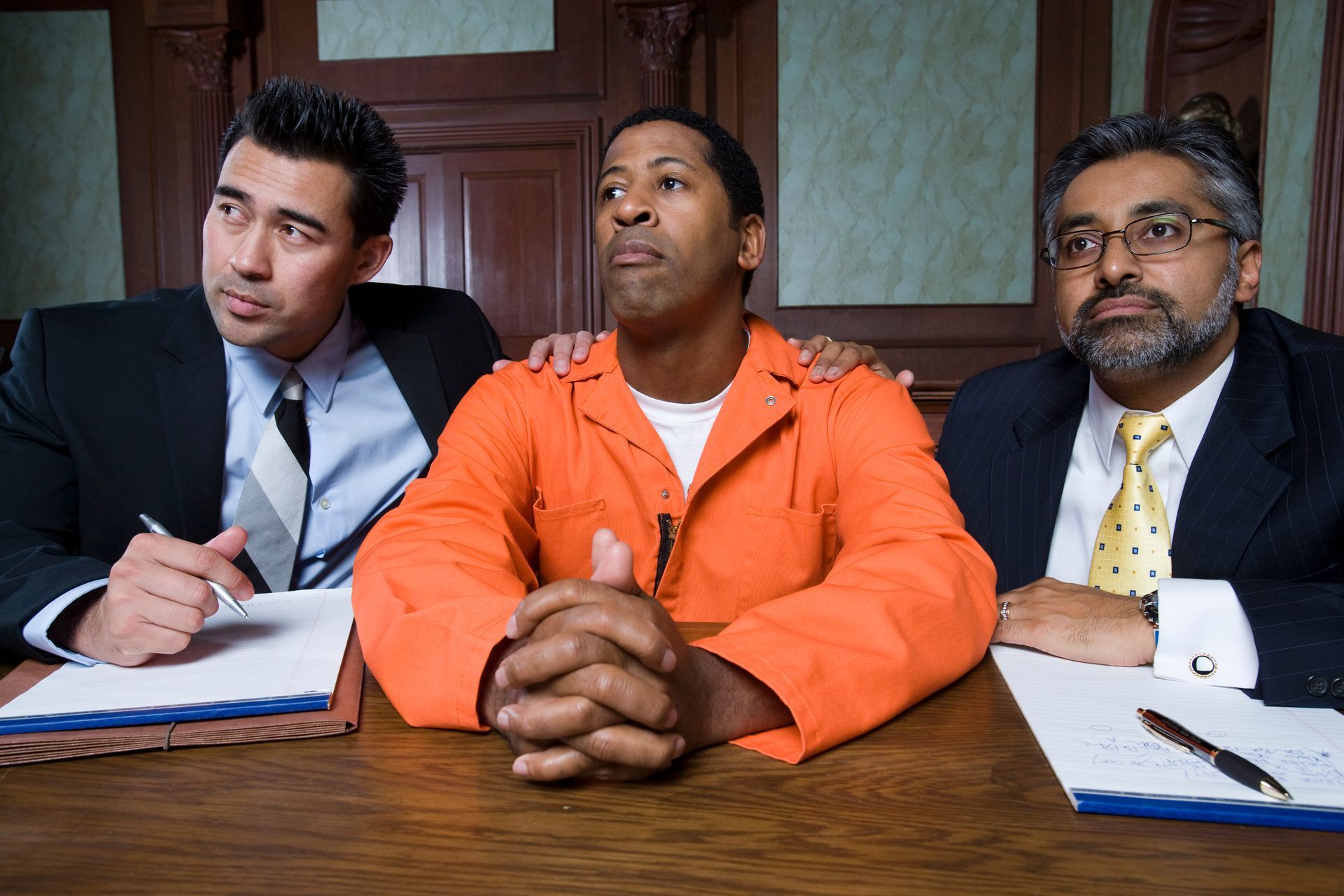 Man in orange jumpsuit flanked by two lawyers in courtroom.