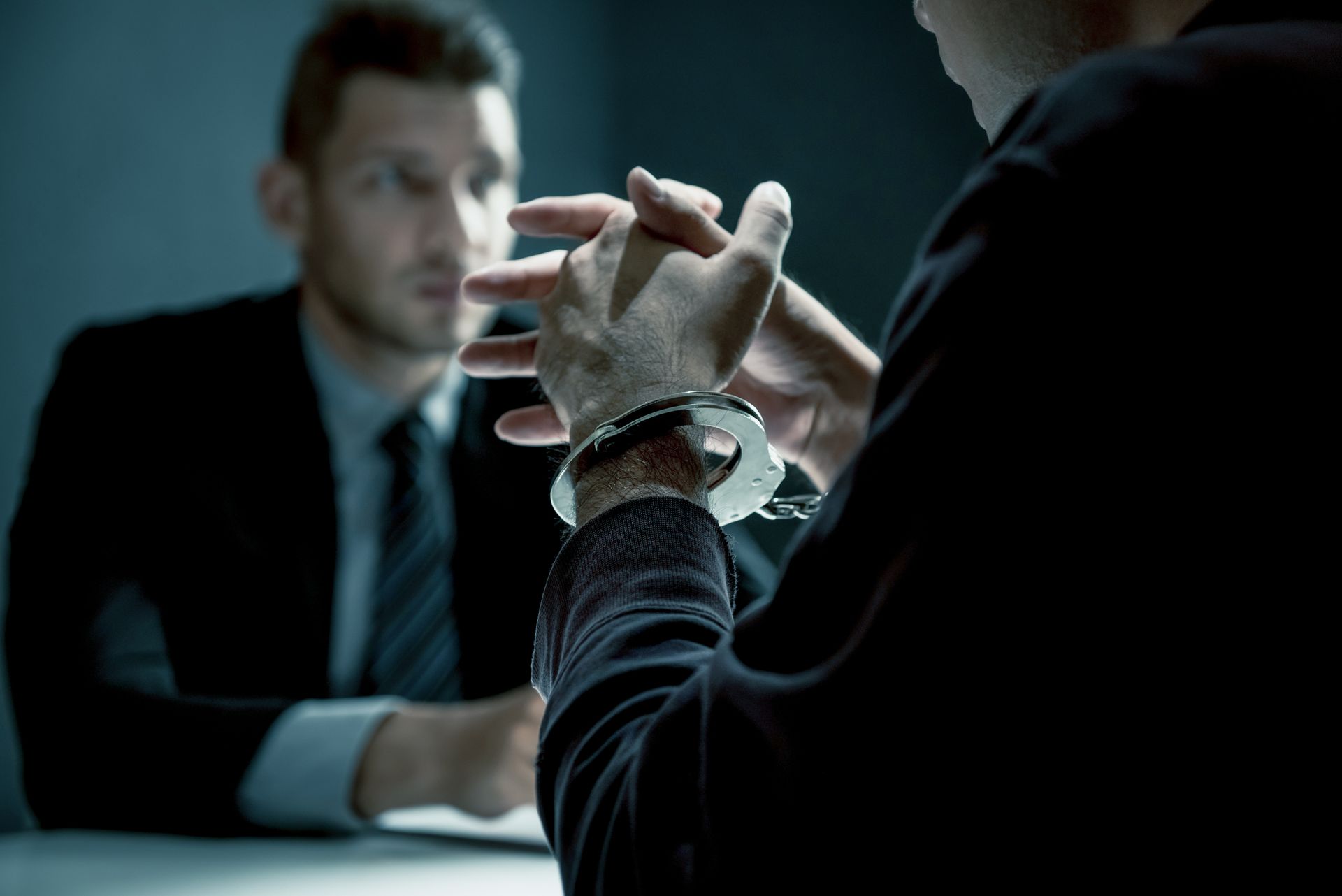 Person in handcuffs at a table, being interviewed by another person wearing a suit.