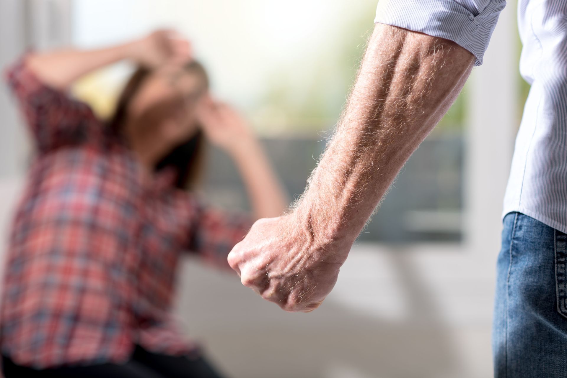 Man's fist raised towards woman in blurred background. She raises hands in fear. Inside by window.