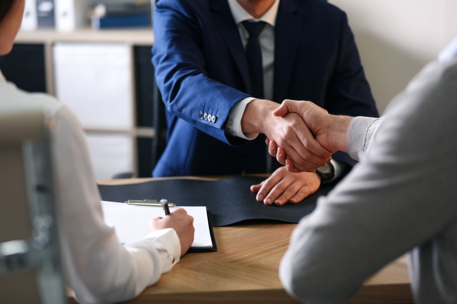 Two men shaking hands across a desk as a person writes. Business setting with neutral colors.