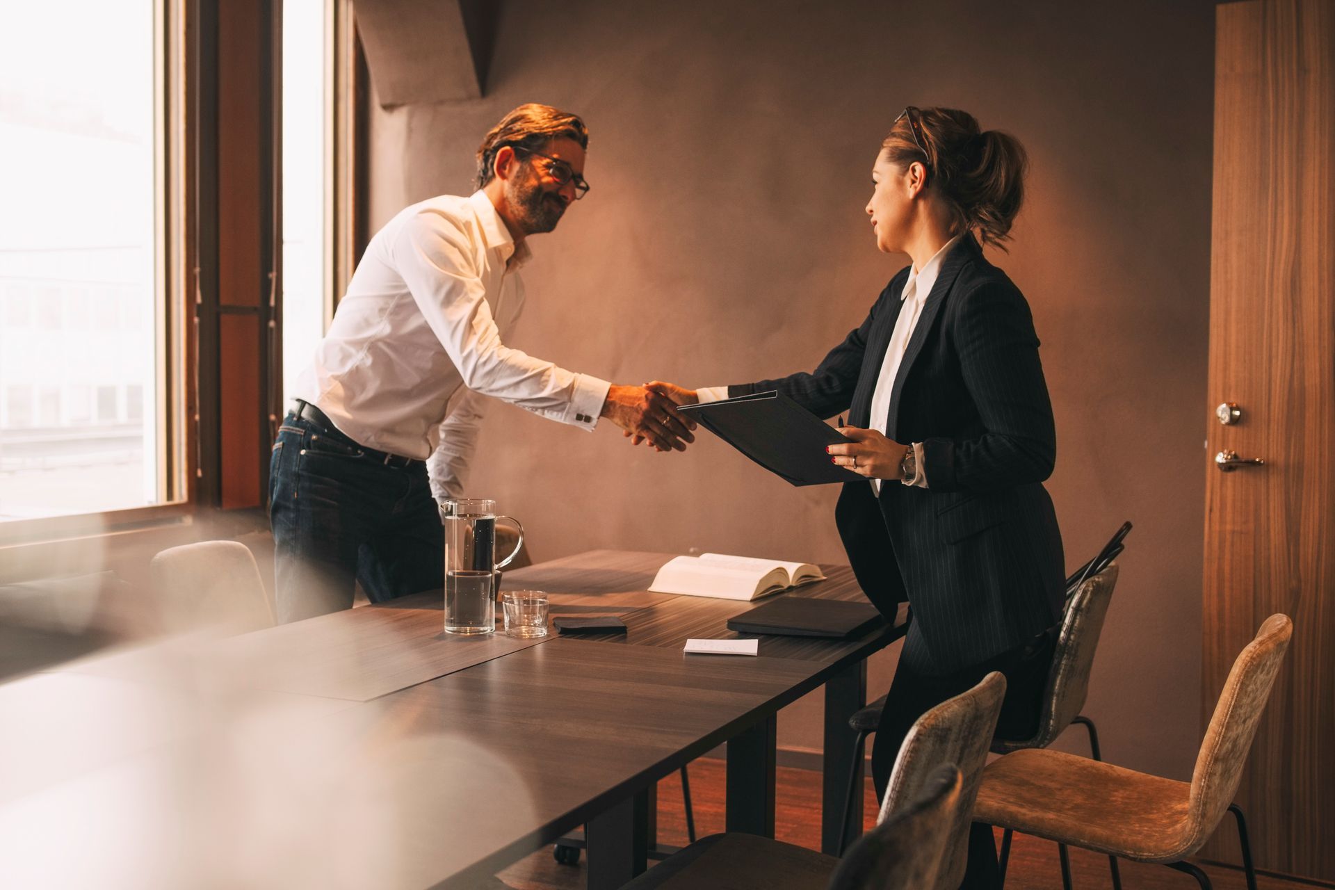 Man and woman shaking hands across a conference table, smiling. Bright room with window.