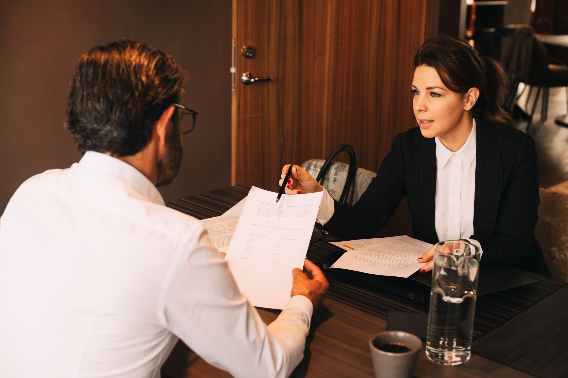 Woman in blazer pointing at a document, interviewing a man in white shirt, seated at a table.