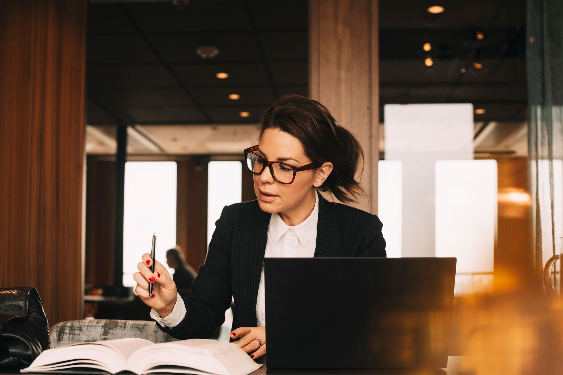 Woman wearing glasses, blazer, reading book and using laptop in a modern office setting.