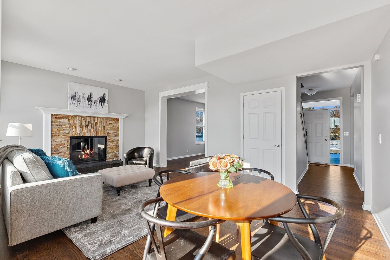 Living room with fireplace, dining table, and entryway. Gray walls, light brown flooring, and natural light.