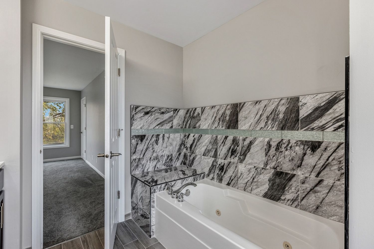 Bathroom with gray and white marble tile and a white jacuzzi tub. A door opens to a hallway with a window.