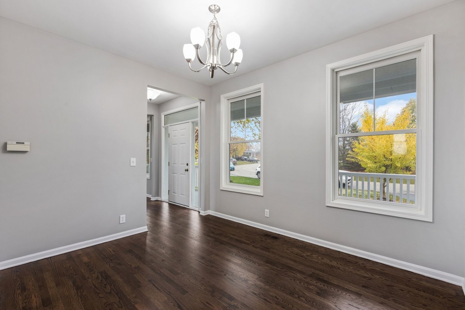 Empty dining room with gray walls, dark wood floors, and chandelier. Two windows provide natural light.