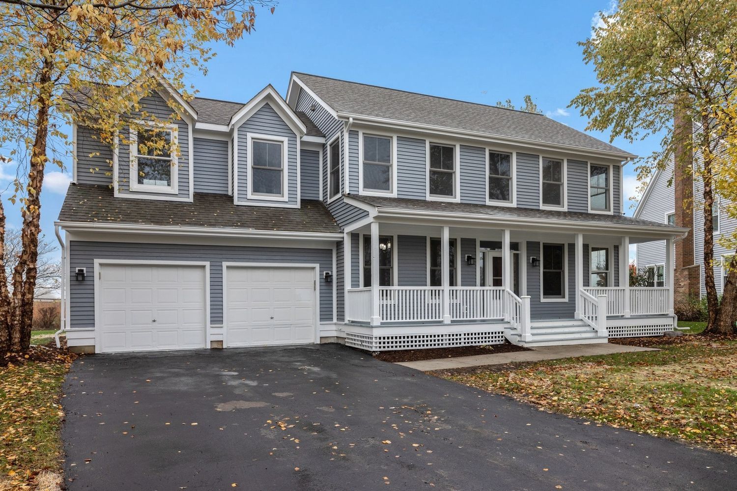 Two-story blue house with a porch and a two-car garage. Autumn trees frame the asphalt driveway.