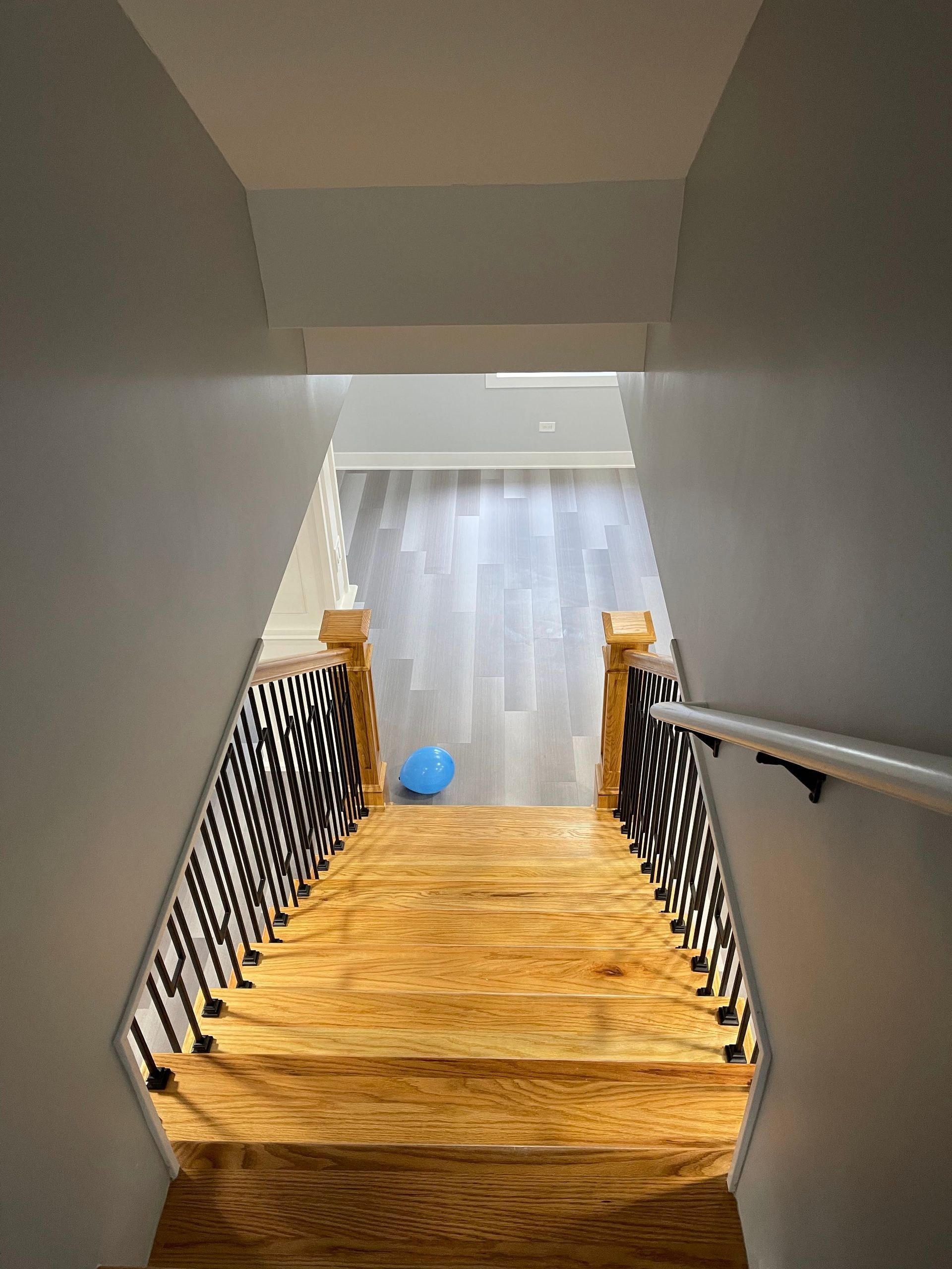 Staircase leading down with wood steps and black railing, ending at a light wood floor with a blue balloon.