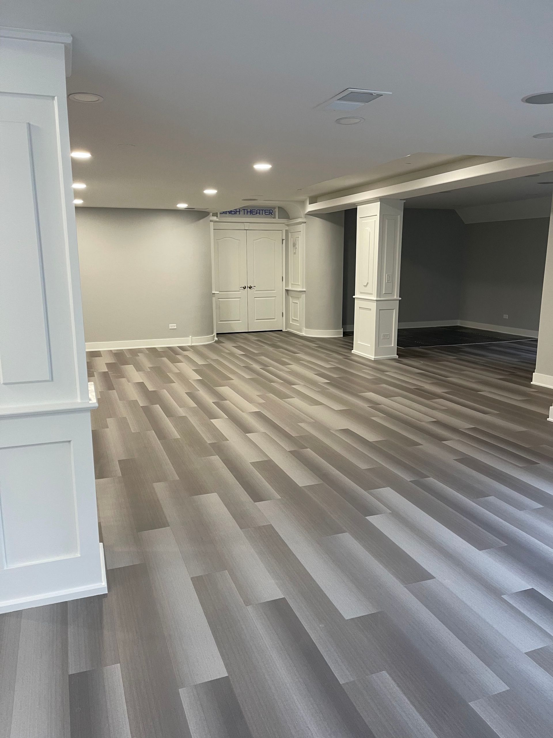 Empty, gray-floored room with white accents, recessed lighting, and a door; likely a finished basement.