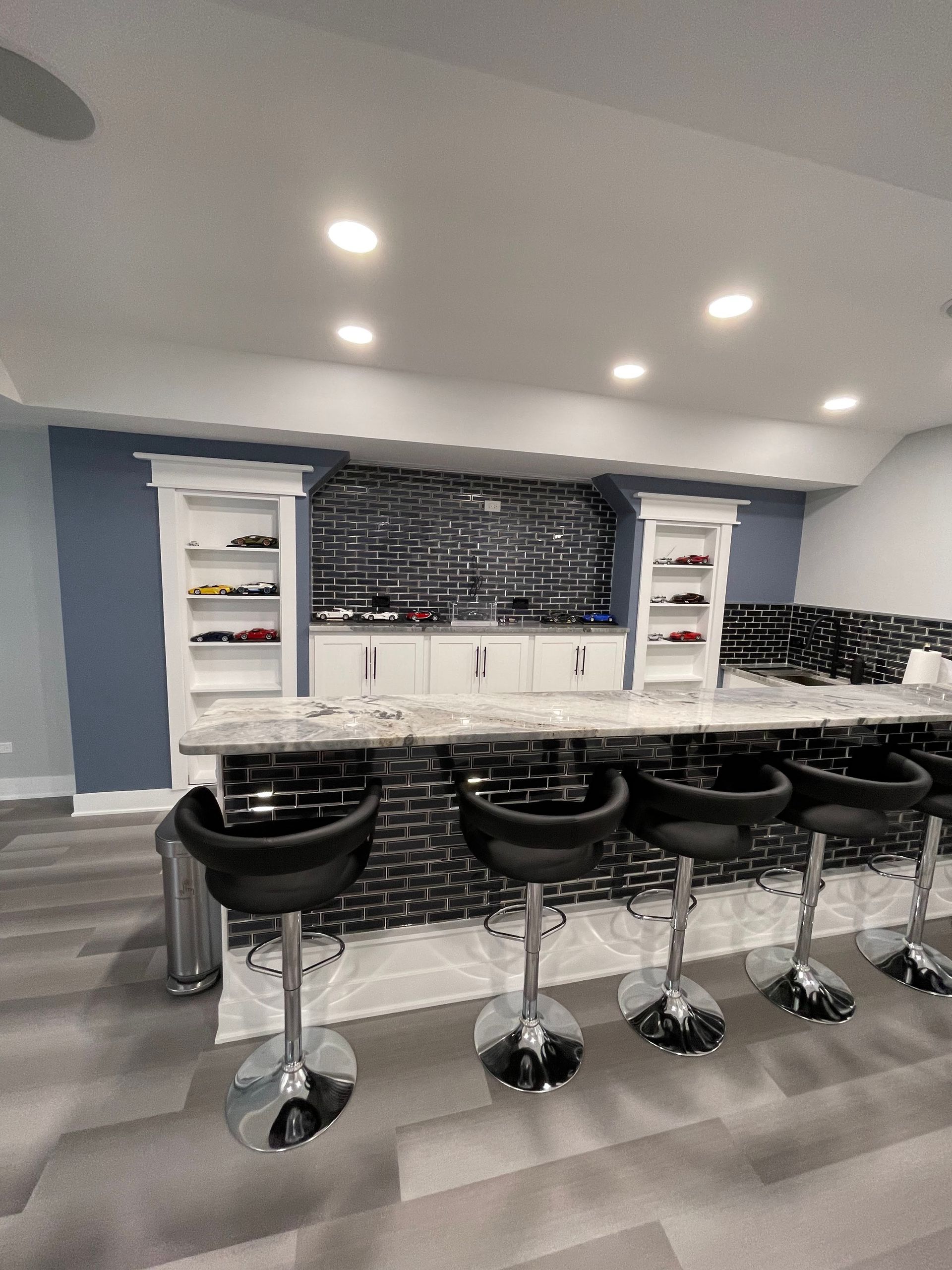 Basement bar with black and white accents. Four black bar stools, built-in shelving, and tile backsplash.
