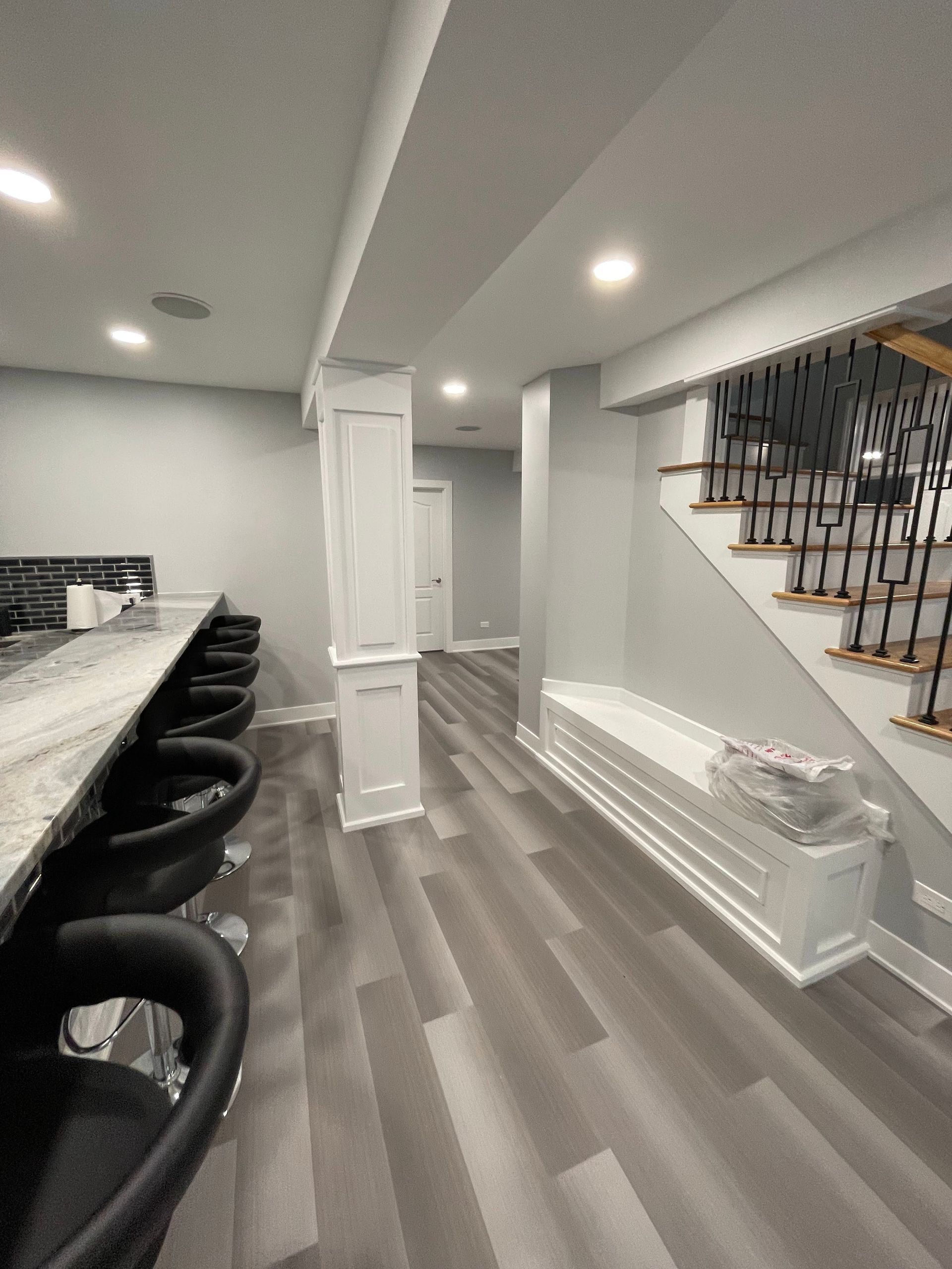 A finished basement with a bar, seating, and stairs with dark metal railing. Gray flooring and walls.