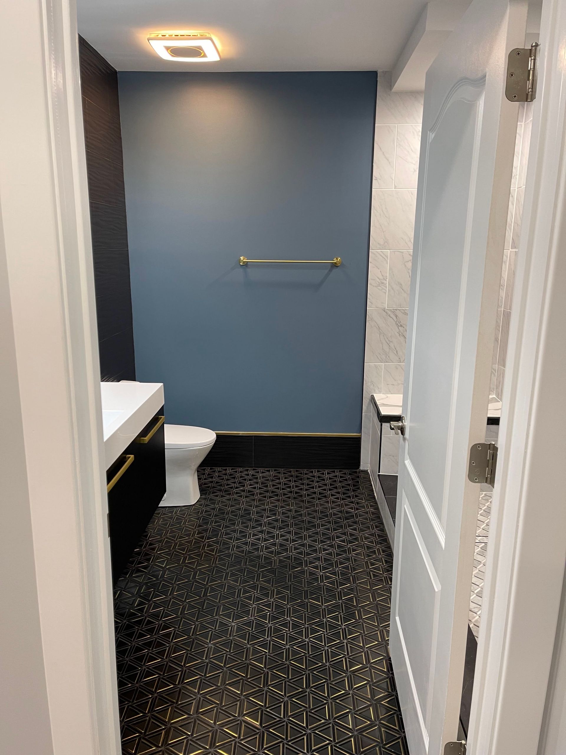 Narrow bathroom with black and gold patterned floor, blue wall, white toilet, and open white door.
