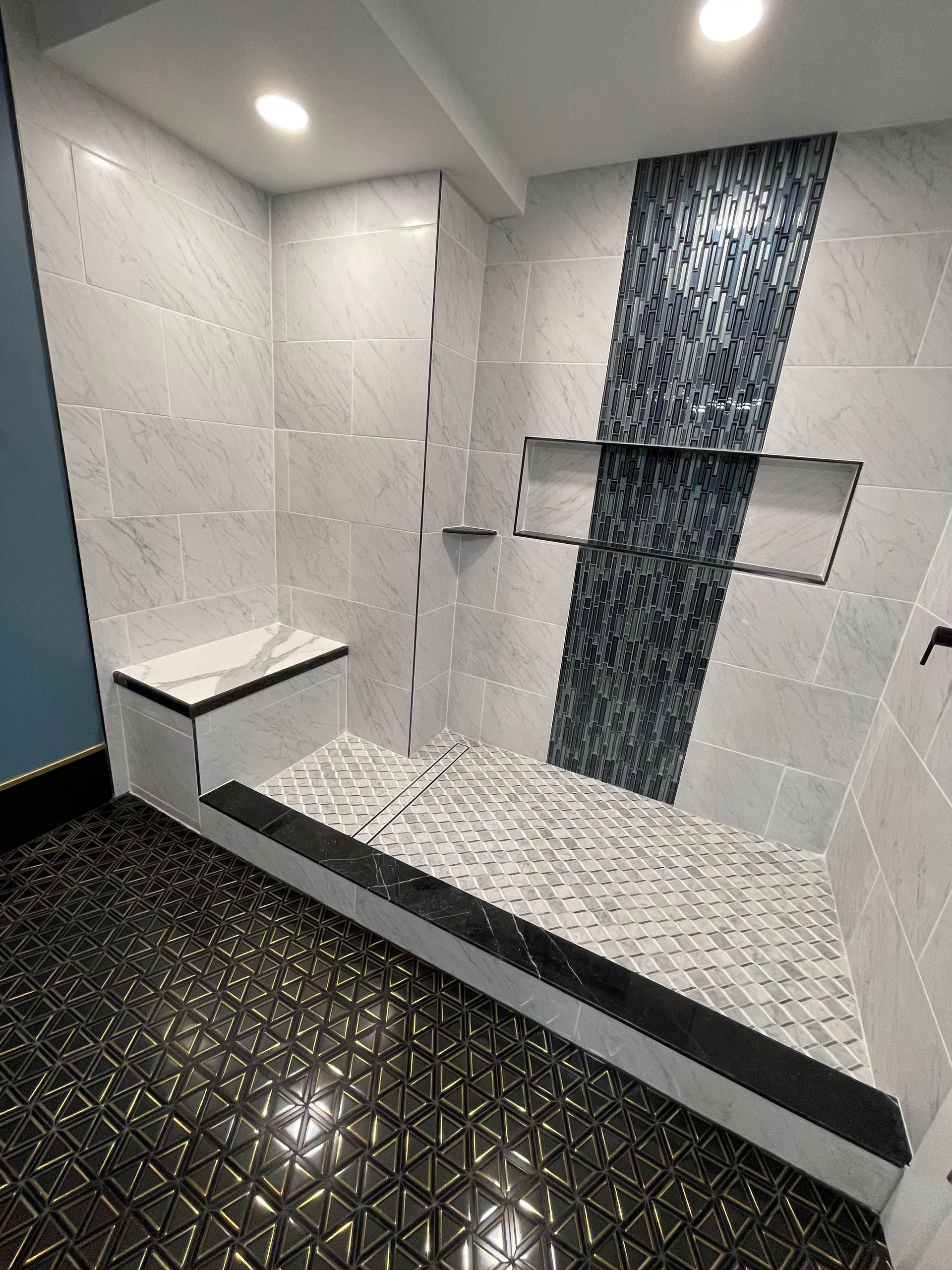 A tiled shower with a black, patterned floor, a white marble bench, and a blue mosaic accent.