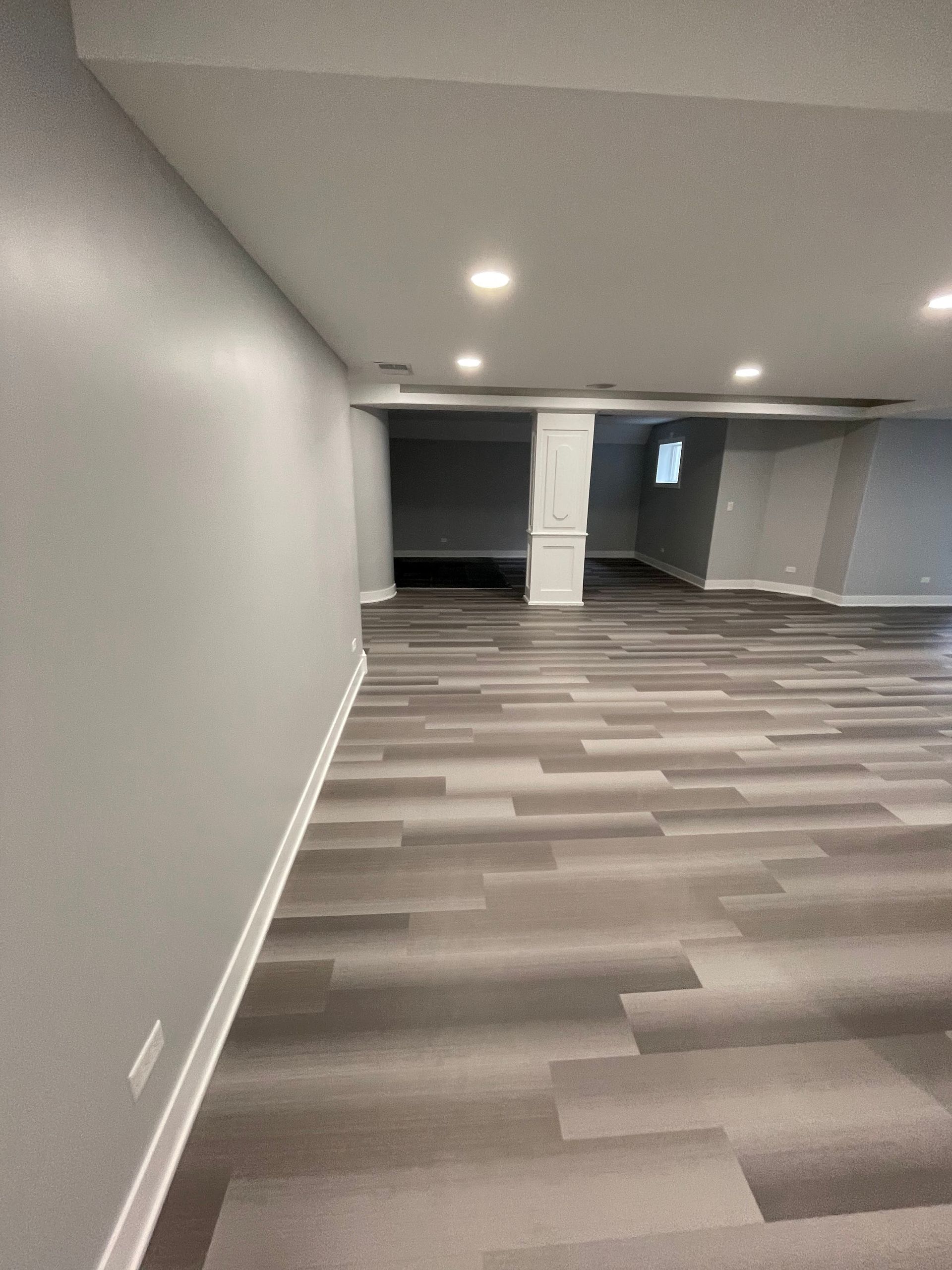 Gray and white finished basement with wood-look flooring. Recessed lighting, a support column, and open doorway.