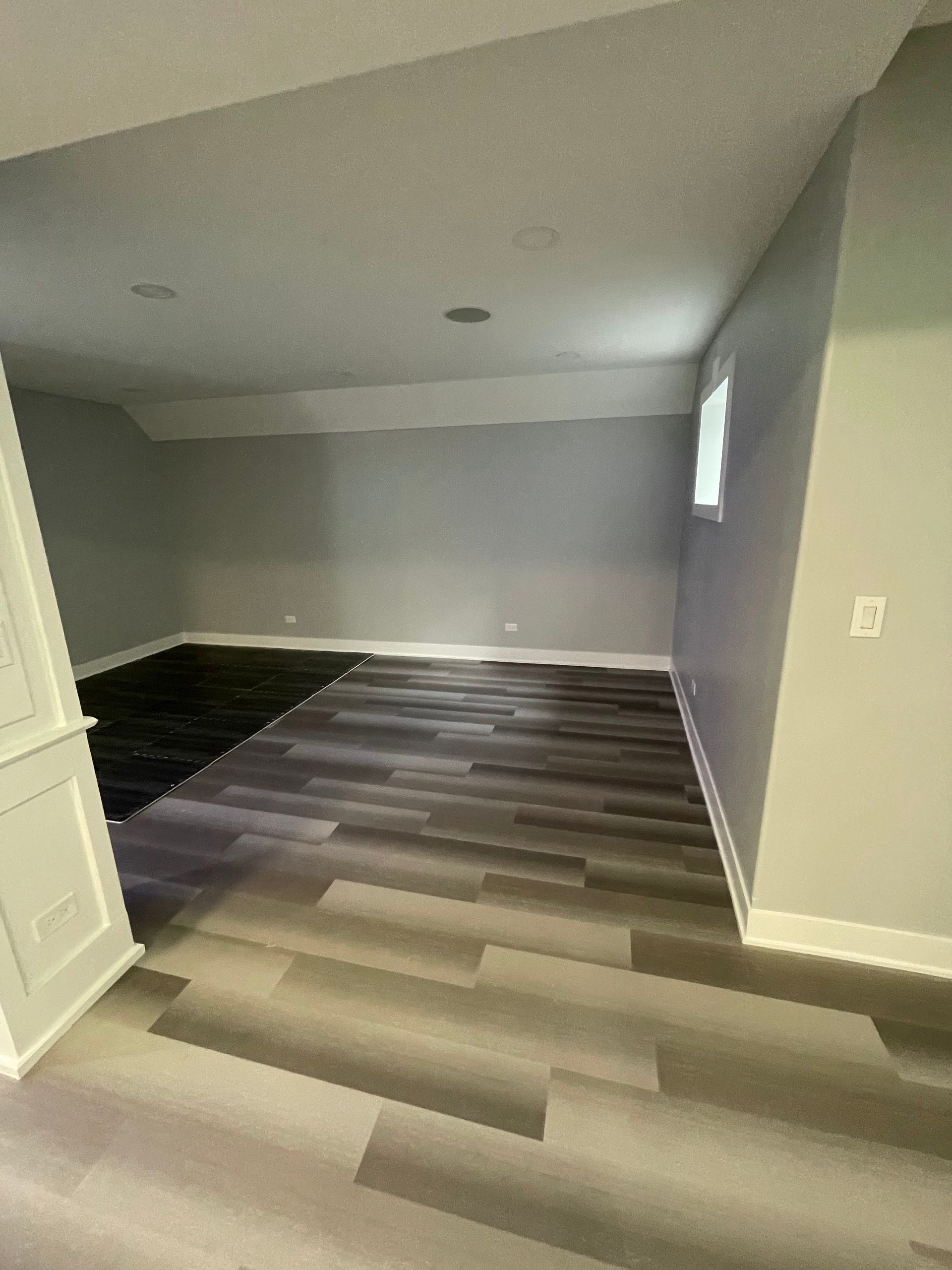Empty room with grey walls and wood-look floor. A black rug and small window are visible.