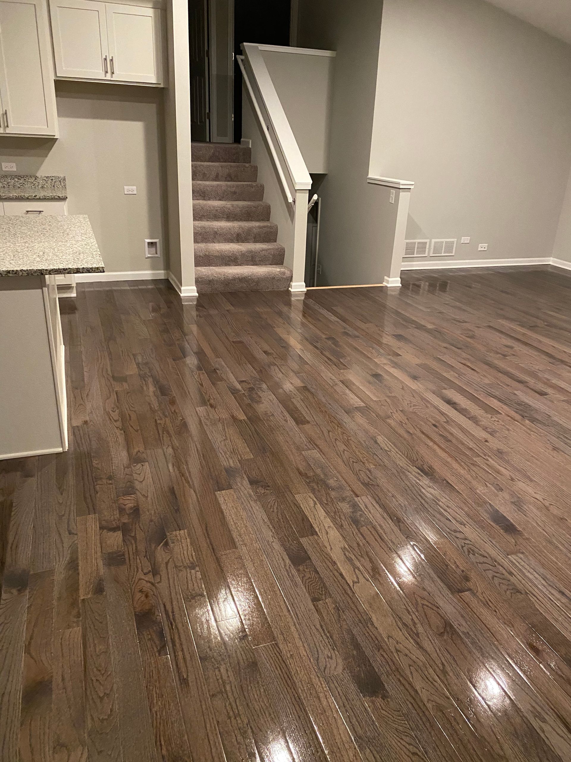 Hardwood floor reflecting light; kitchen cabinets and staircase in background.