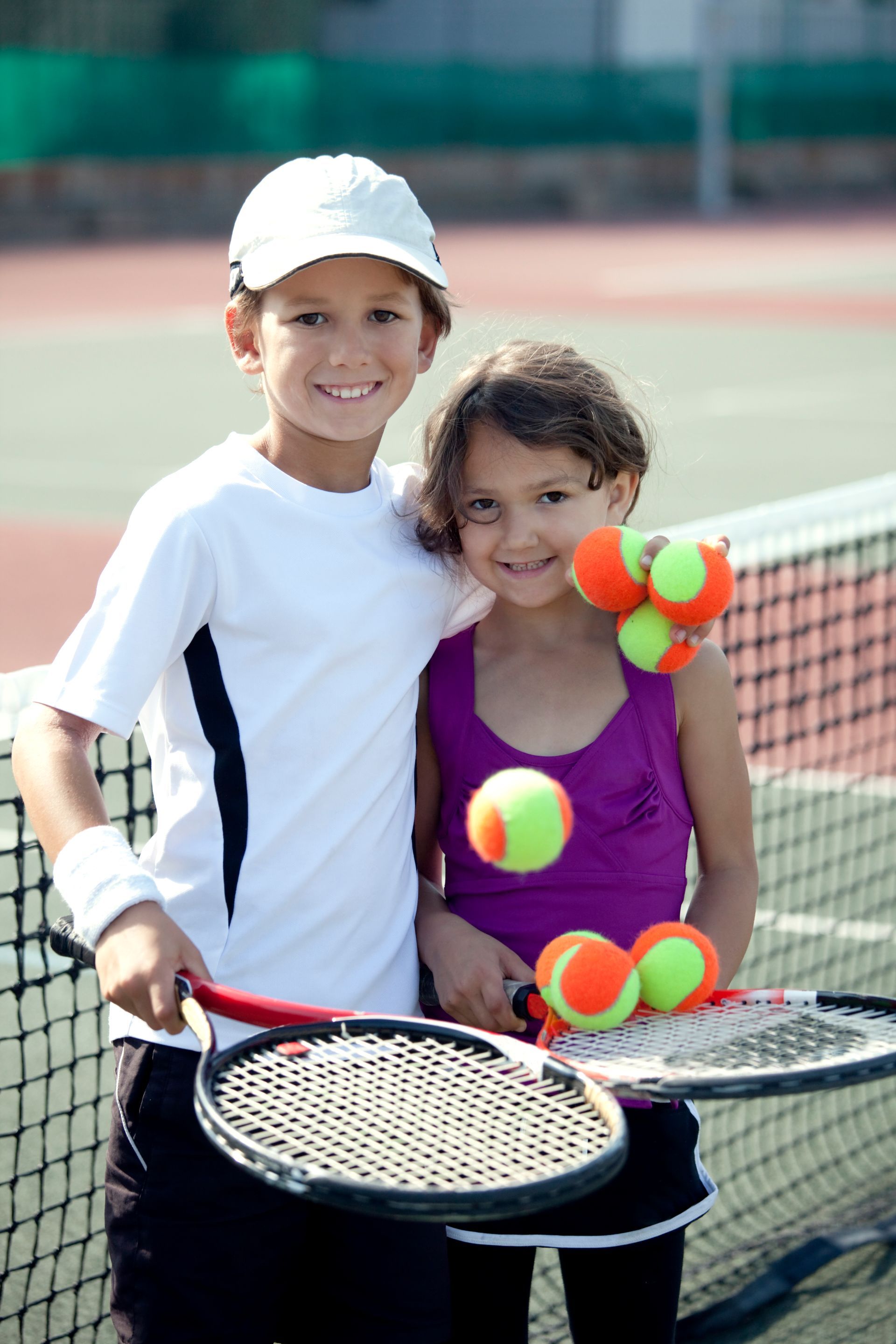 Two tennis players smiling on a court, holding rackets and tennis balls beside the net