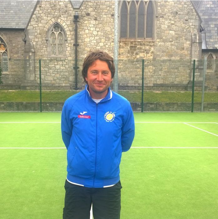 Man in blue jacket standing on green field in front of stone building and fence