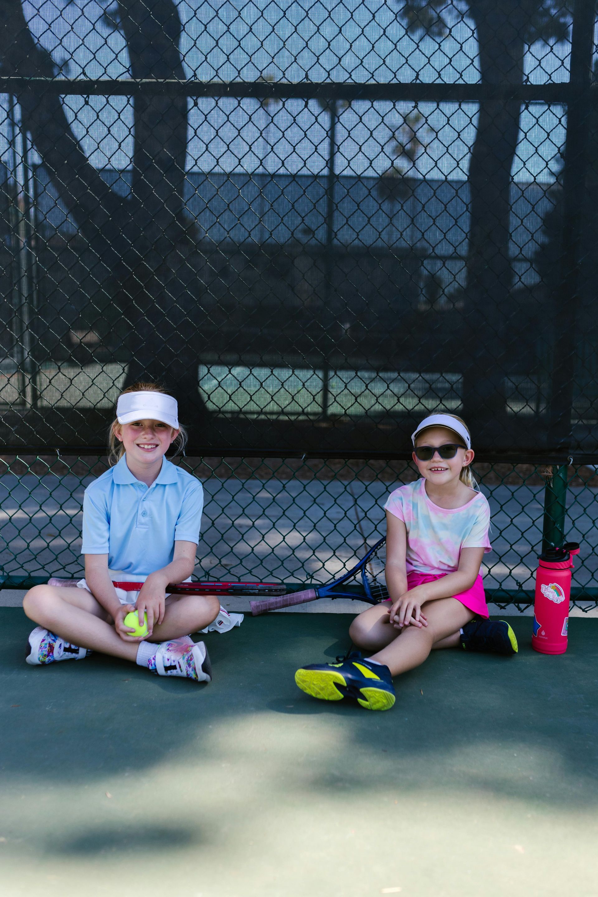 Two children sitting on a tennis court with rackets, balls, and water bottles by the fence