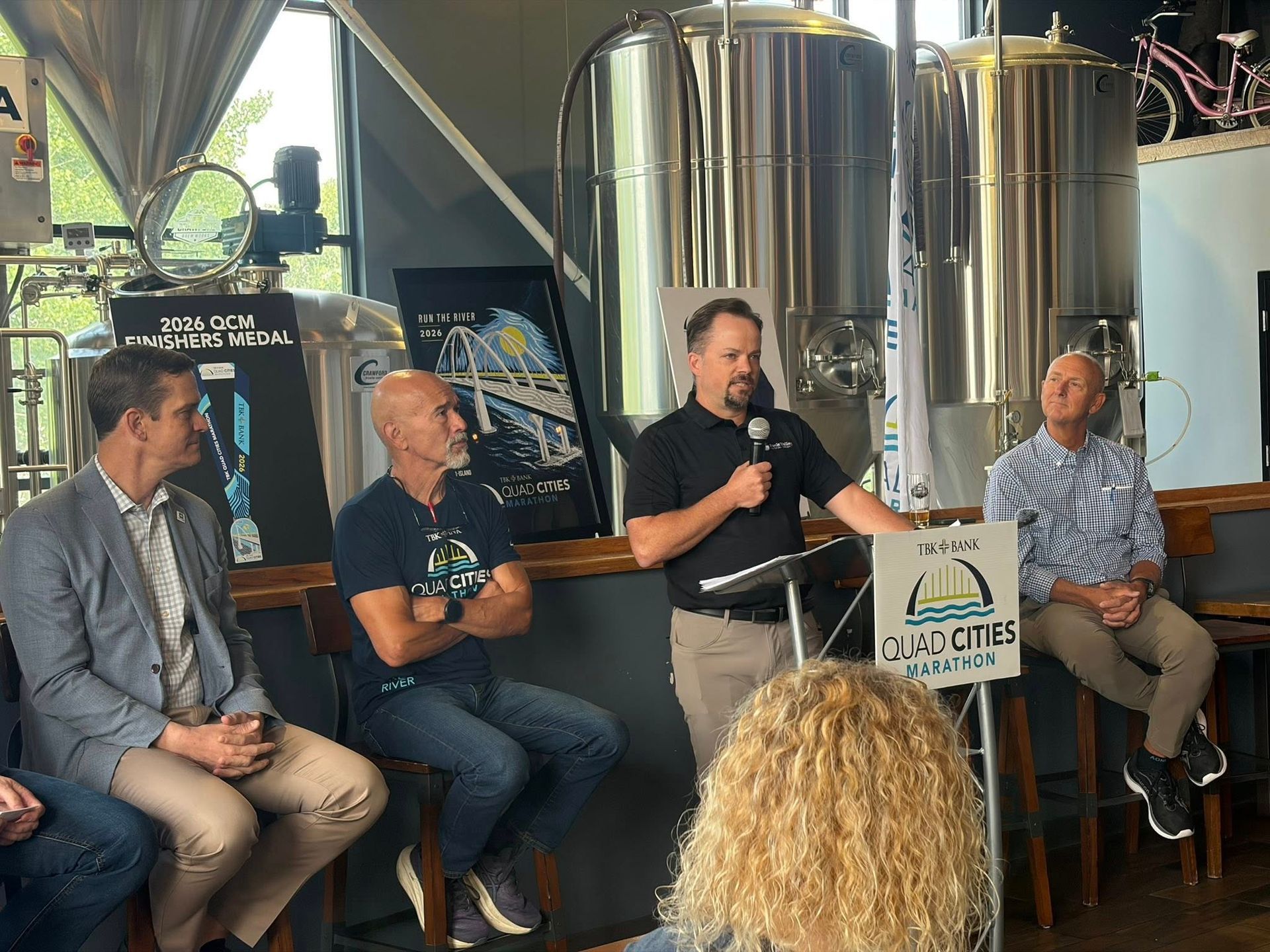 Panel discussion at a brewery with four speakers seated on stools beside large stainless steel tanks
