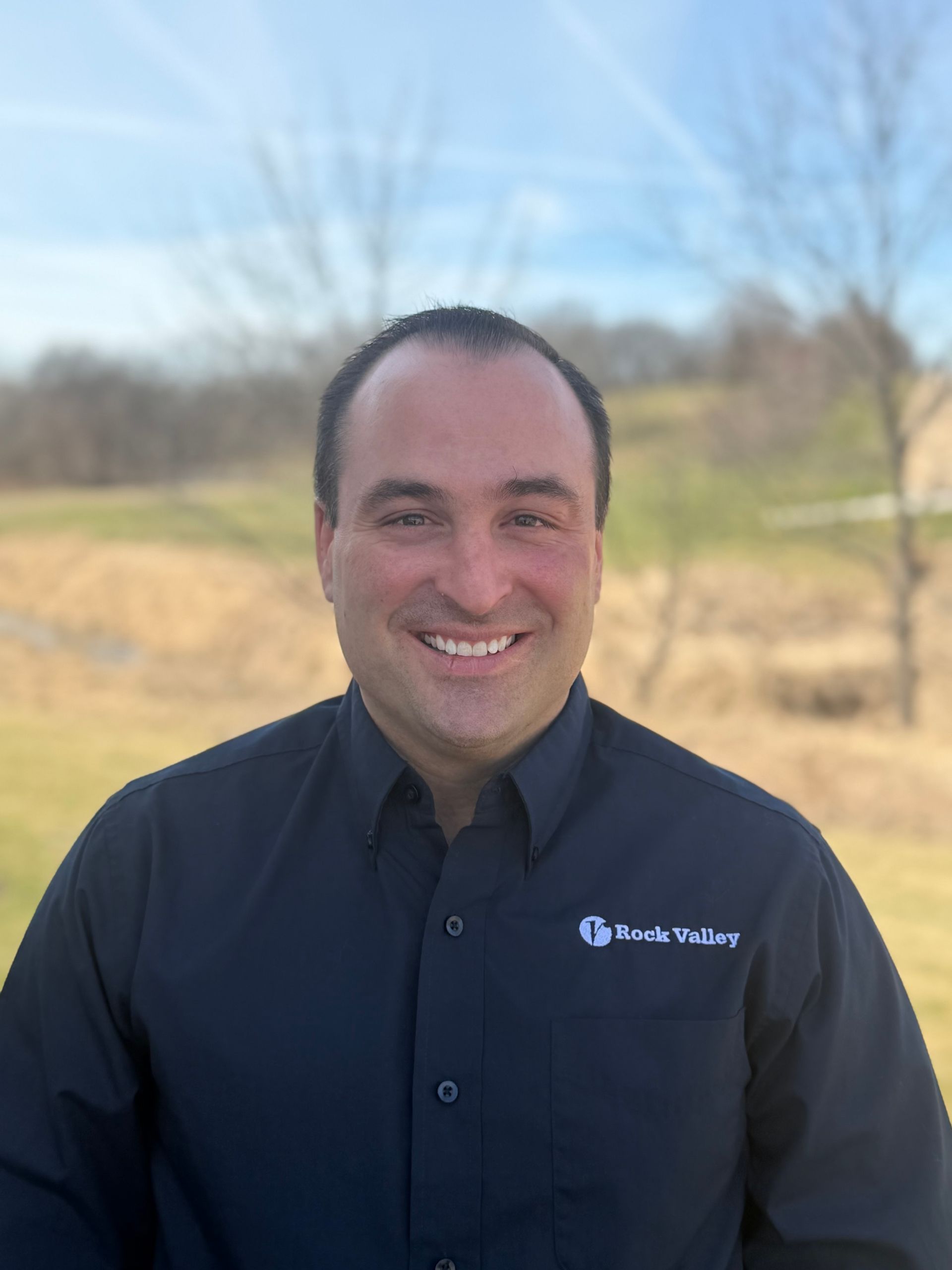 Smiling man in a blue shirt standing in front of a green hedge