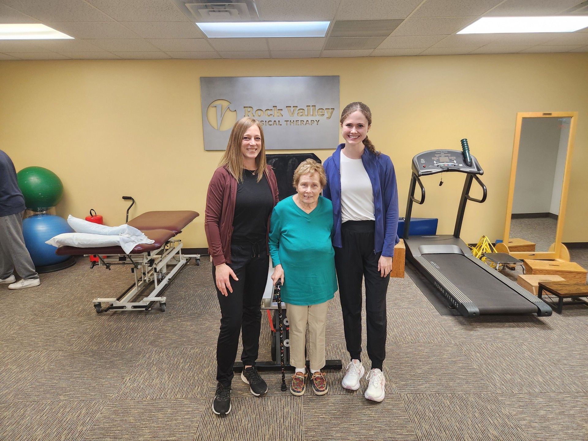 Three people stand in a therapy gym with treadmills, exercise equipment, and a mirror.