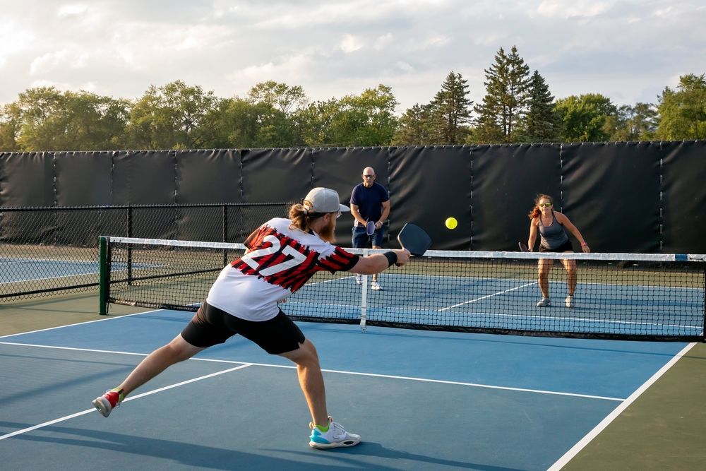 Pickleball players rally on an outdoor court, with a player in a white jersey lunging for the ball.