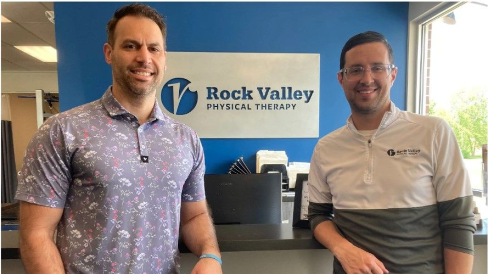 Two men standing at the Rock Valley Physical Therapy reception desk, smiling in a lobby