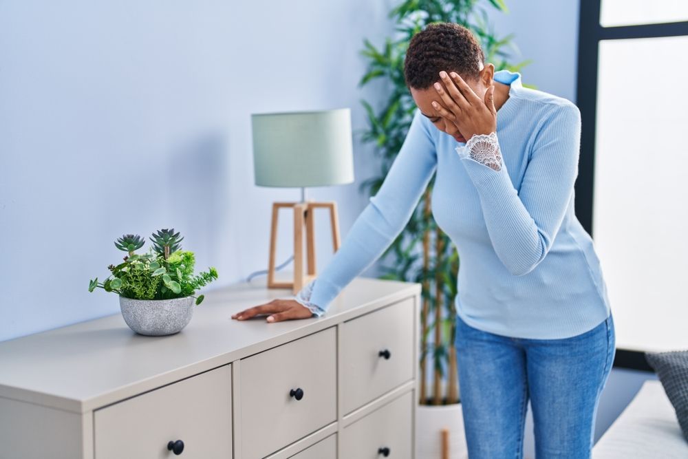 Person in blue sweater stands beside a cabinet, covering their face in a bright room with a plant and lamp.