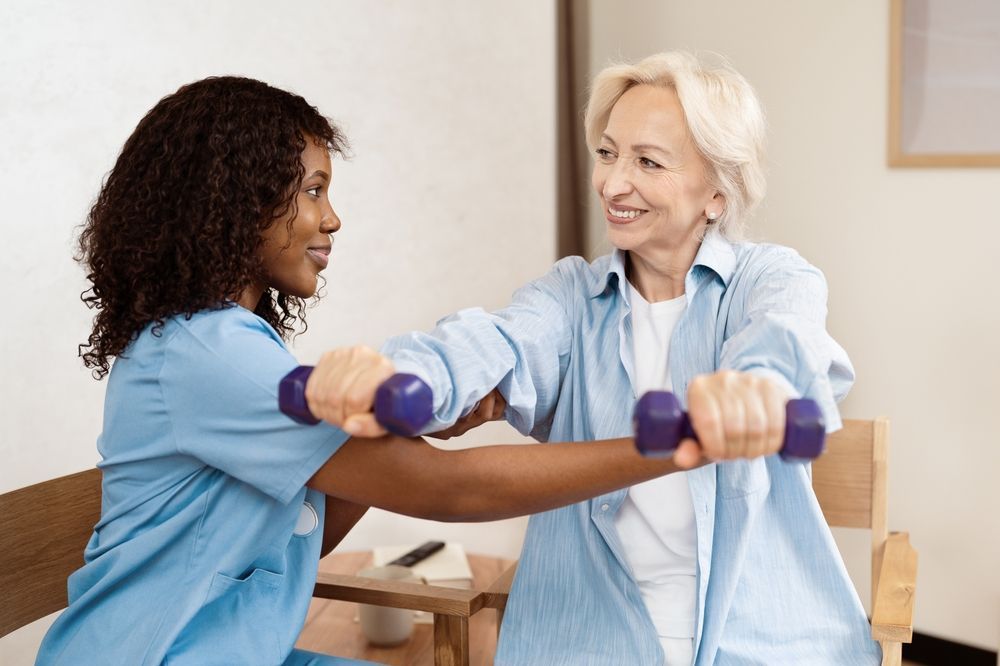Therapist assists a person with exercise, holding dumbbells. Interior, light-colored room.