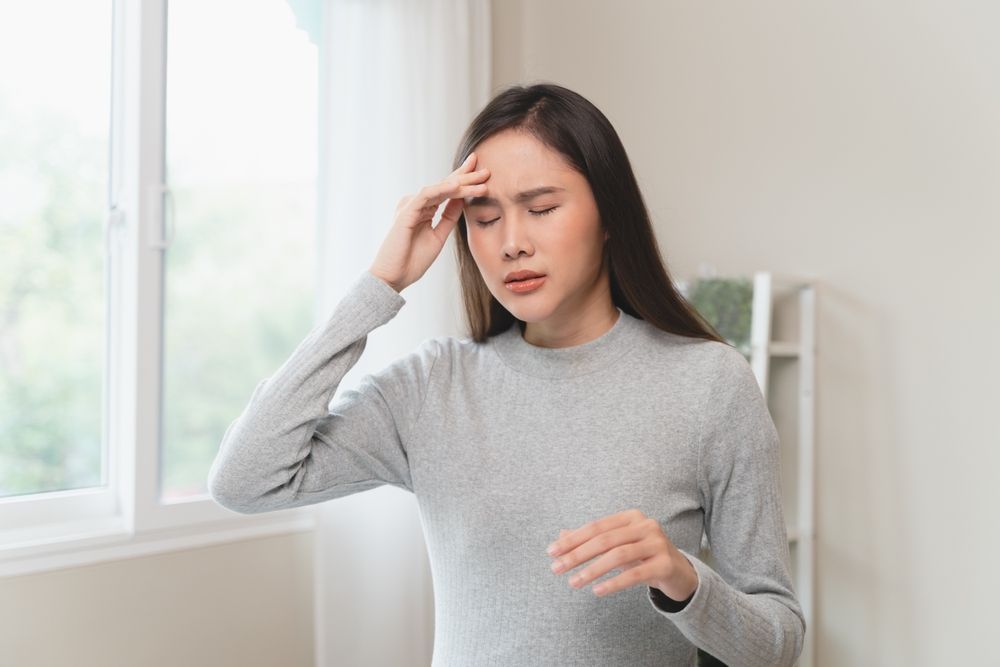 Woman in a gray sweater holding her forehead and looking pained in a bright room by a window