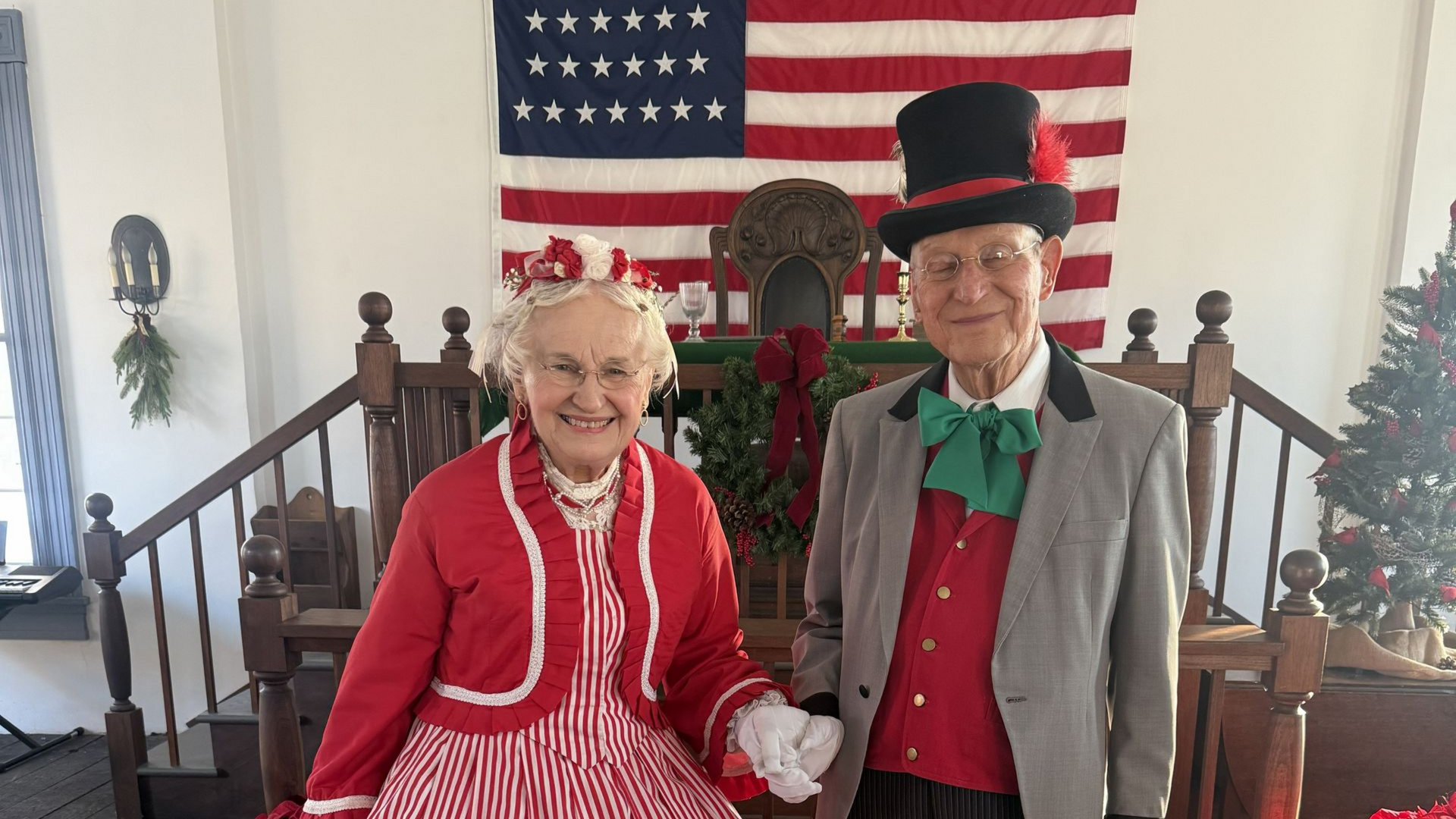 Two people in festive holiday attire standing in front of a large American flag indoors.
