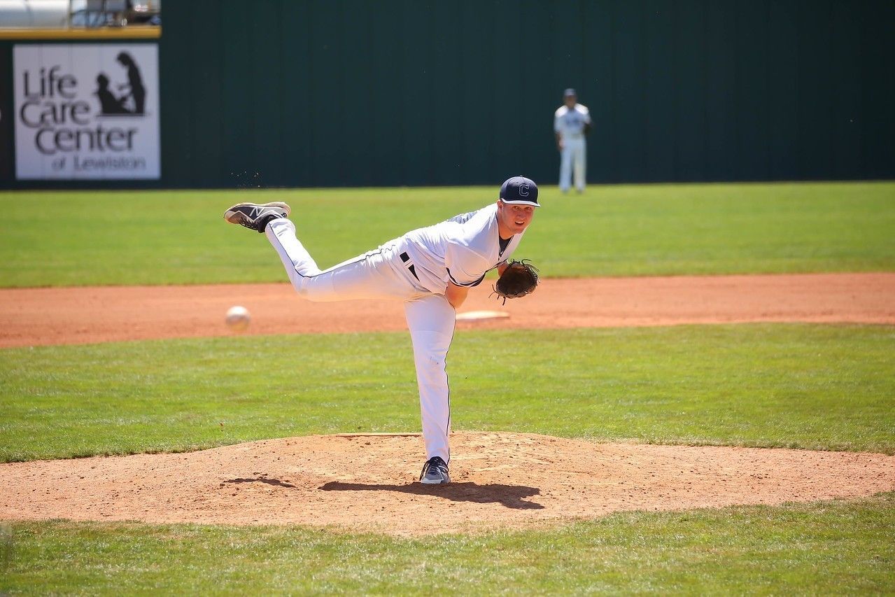Baseball pitcher in white uniform mid-throw on the mound, with outfield wall and player in the background