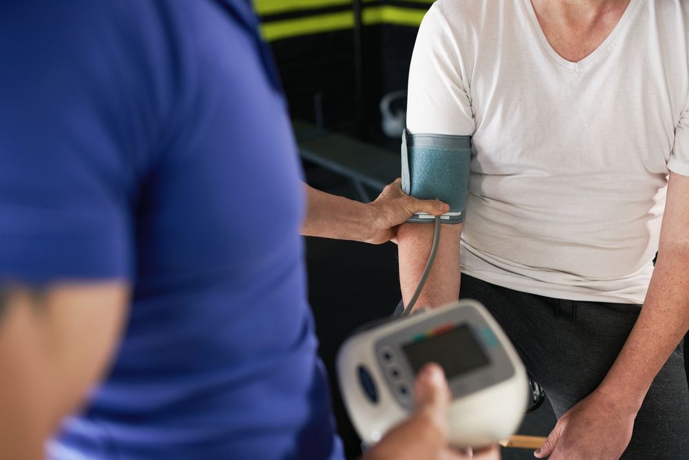 A person in a blue shirt measures the blood pressure of a person in a white shirt using an electronic monitor.