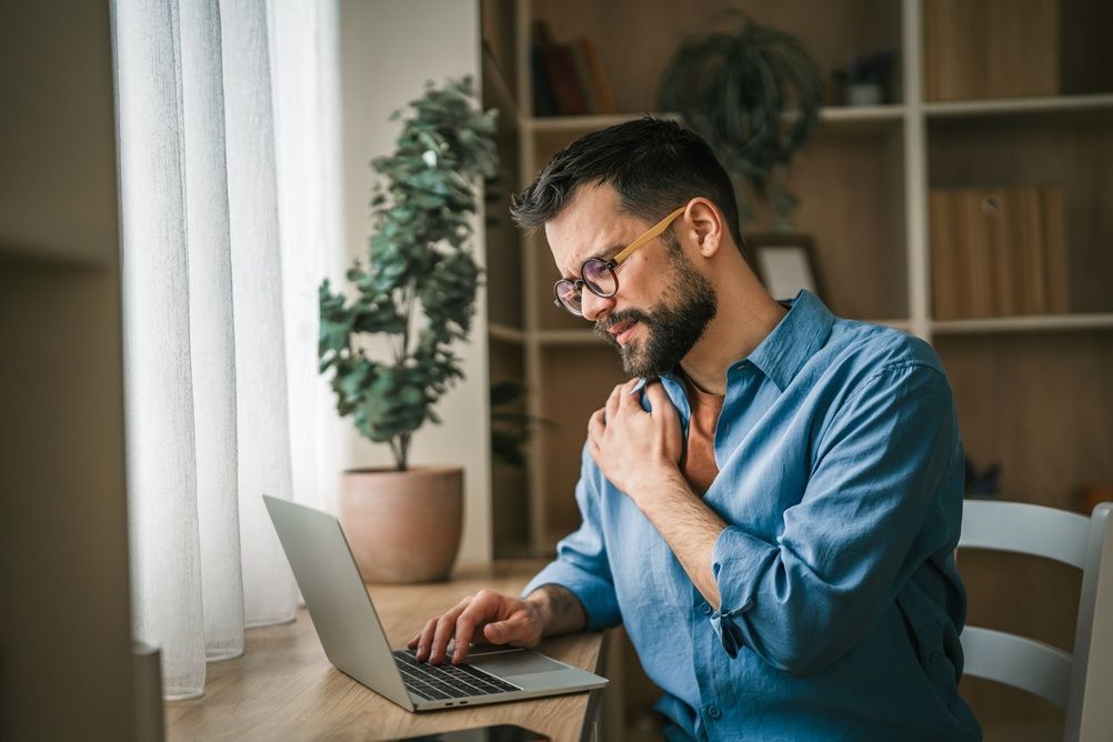 A bearded person in a blue shirt sits at a desk using a laptop, clutching their shoulder with an pained expression.
