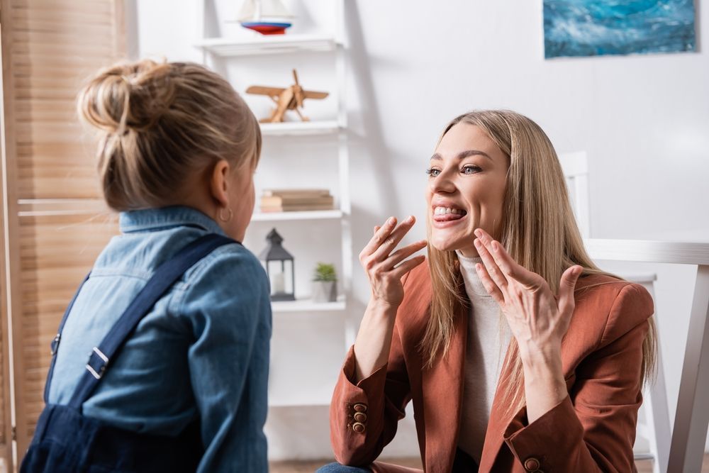 Woman demonstrating mouth movements to a young child, likely a speech therapy session.