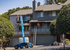 Workers painting a two-story townhouse. One in a lift, one on a balcony, with a ladder and equipment on the ground.