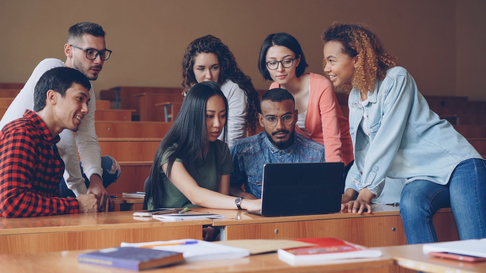 A diverse group of students in a lecture hall looking at a laptop screen together.