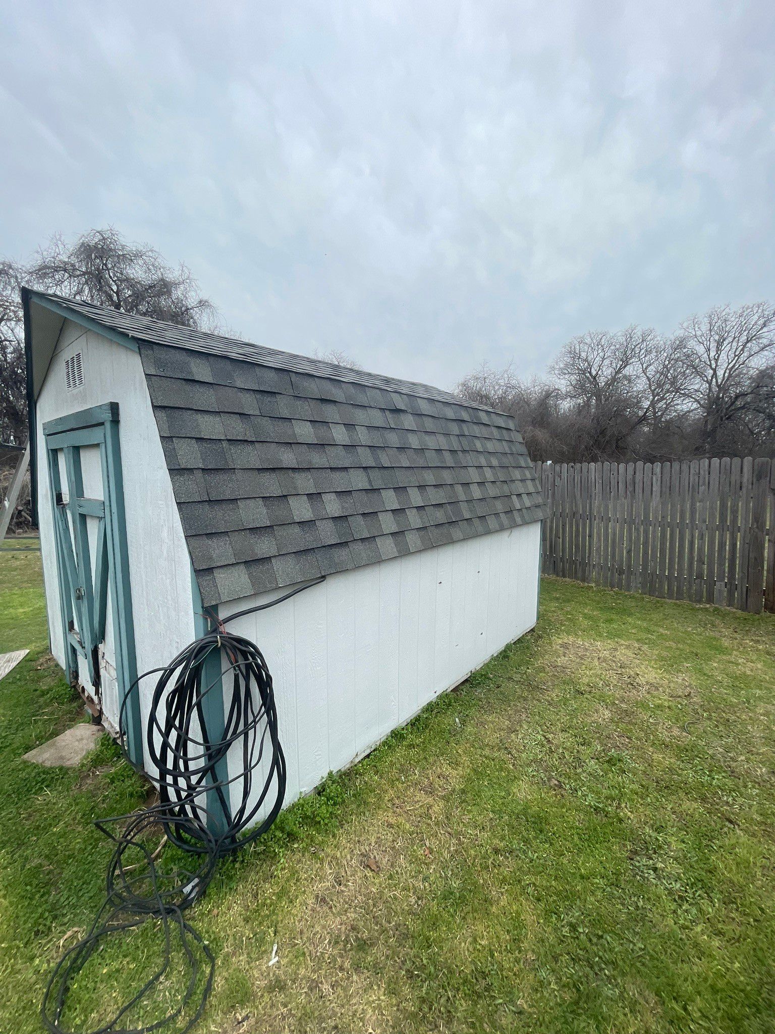 A white shed with a gray roof is sitting in the grass next to a wooden fence.