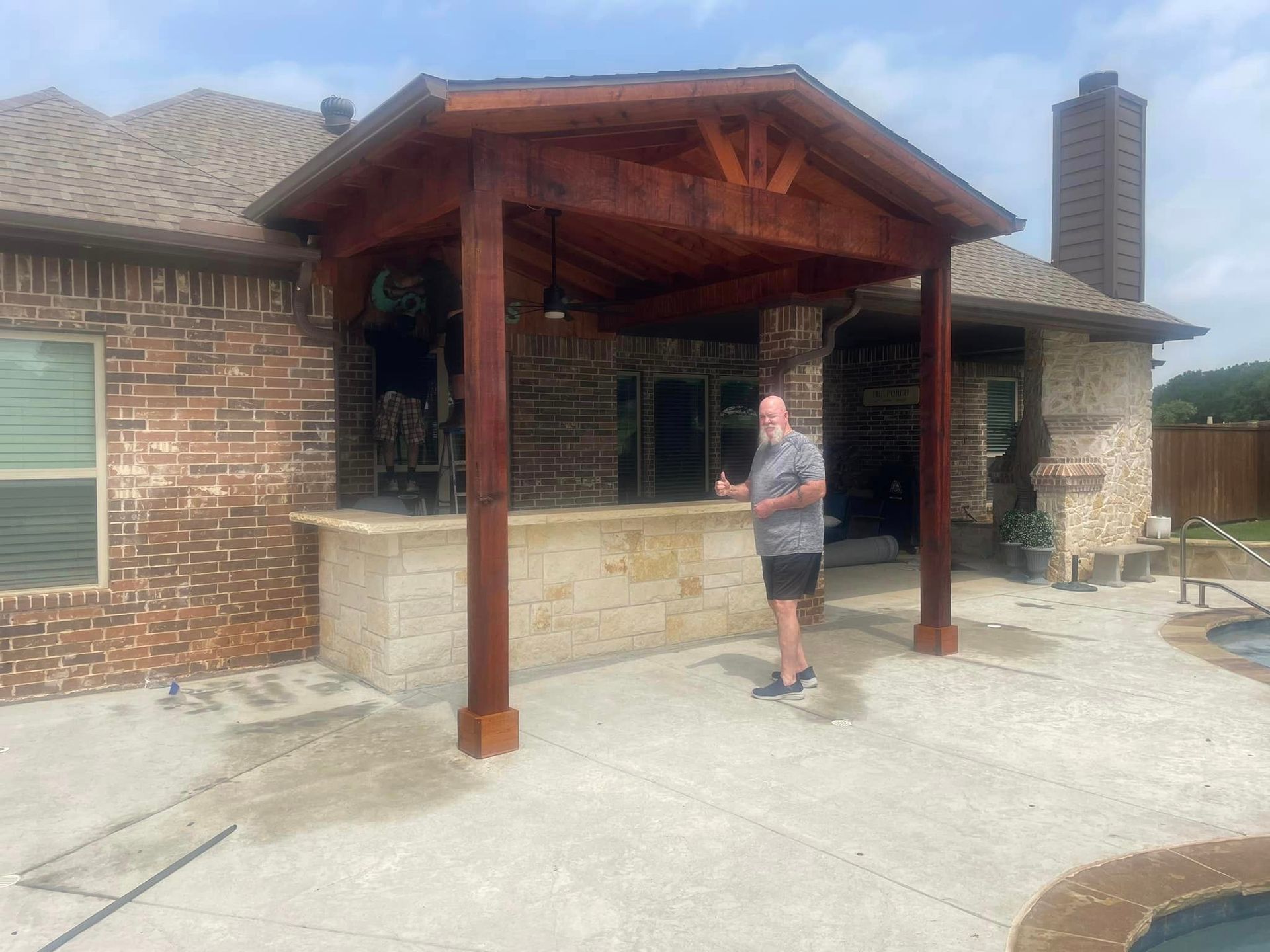 A man is standing under a wooden pergola in front of a brick house.