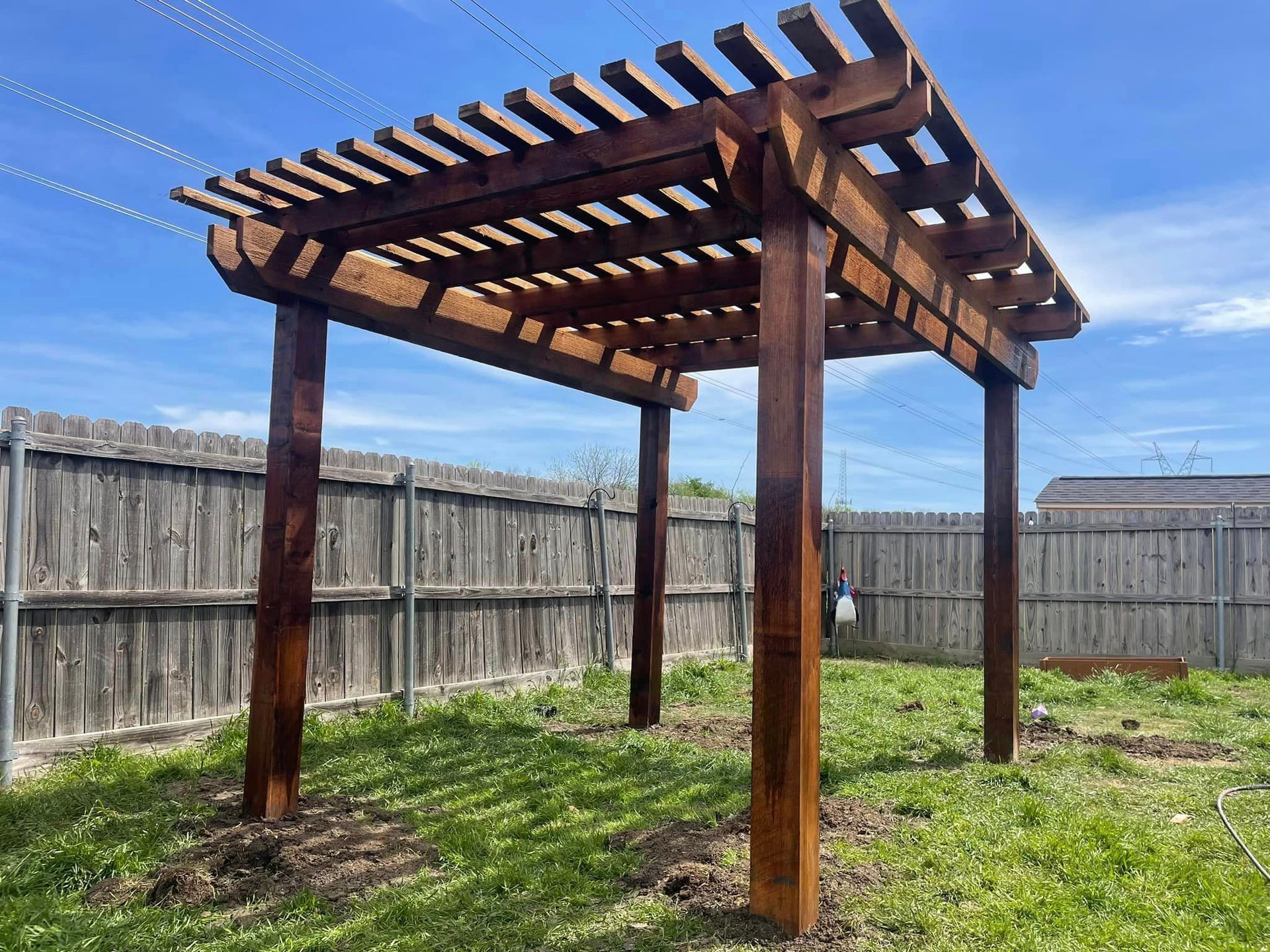 A wooden pergola is sitting in the middle of a grassy yard next to a wooden fence.