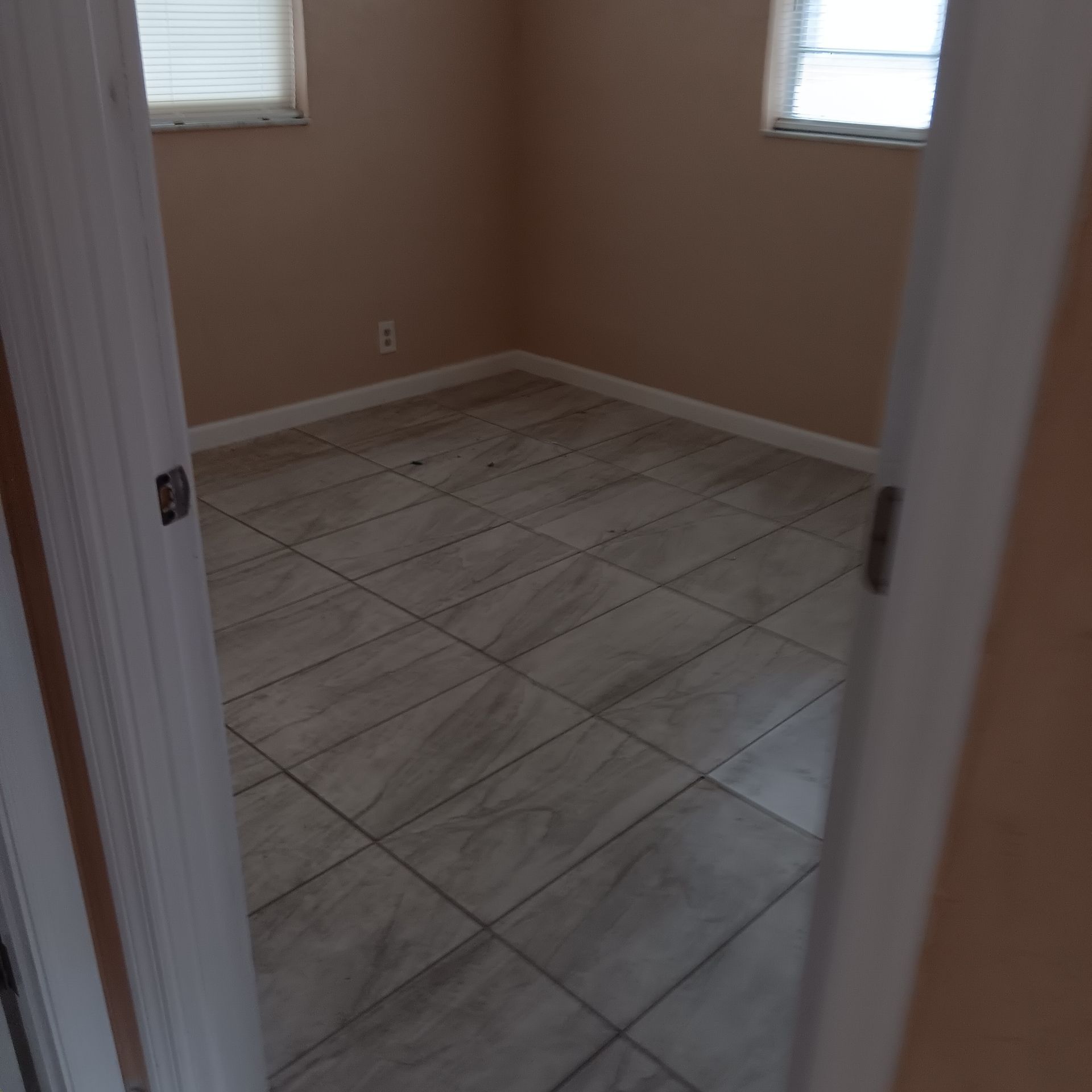 Empty room with beige walls and tile floor, two windows. View through doorway.