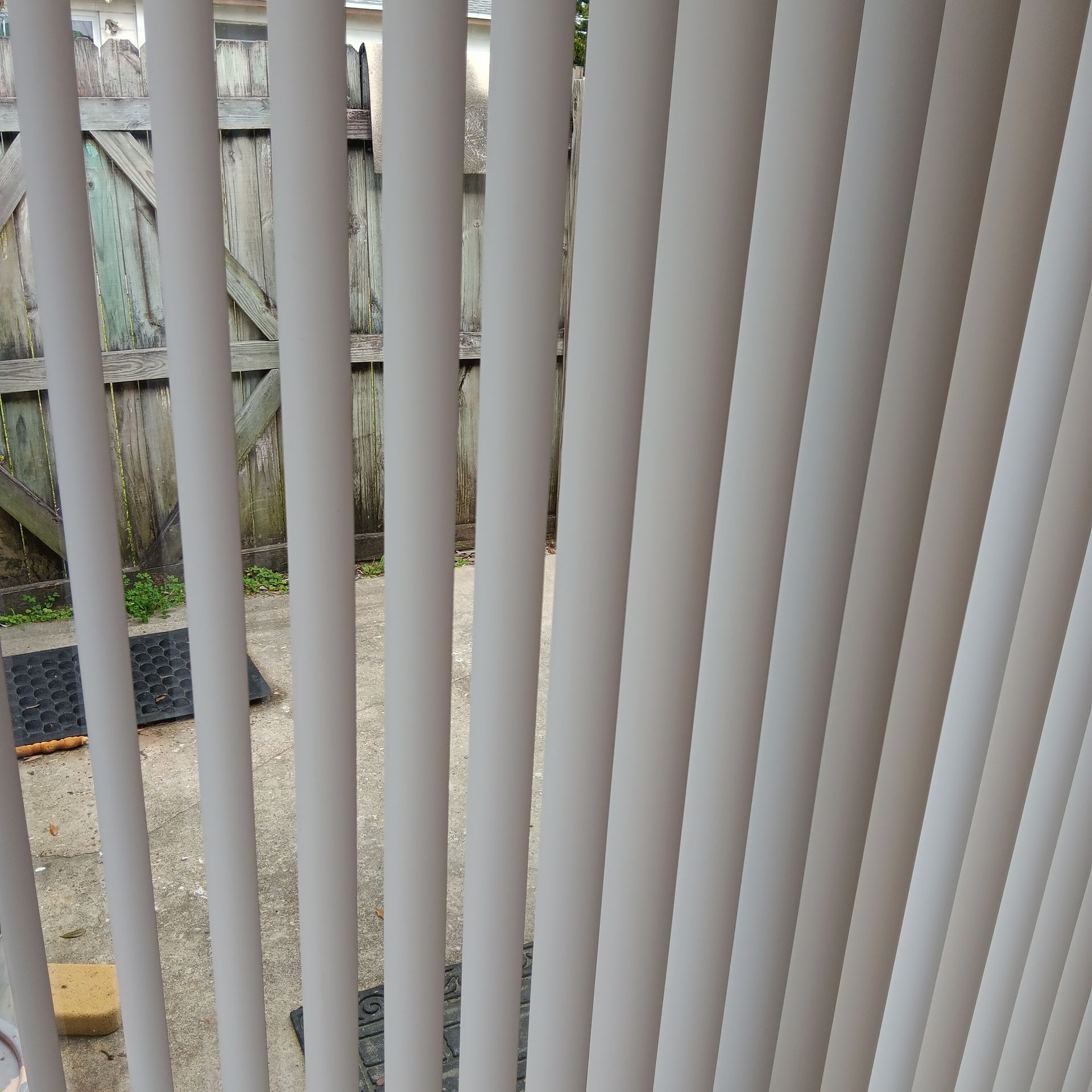 White vertical blinds partially covering a view of a wooden fence and concrete patio.