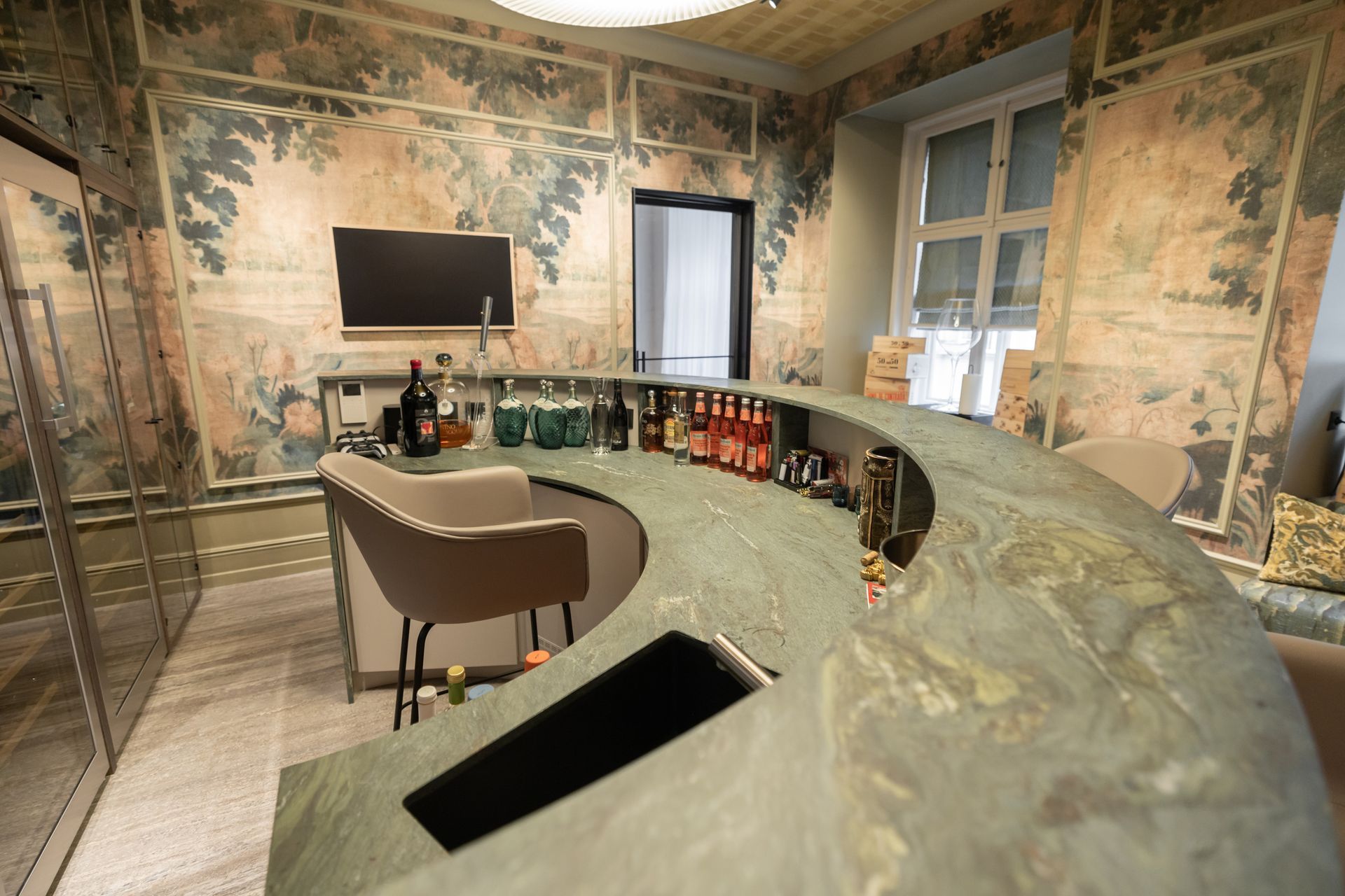 Bar mit geschwungener grüner Theke, gefüllt mit Flaschen. Beiger Hocker, kunstvolle Tapete und Fernseher im Hintergrund.