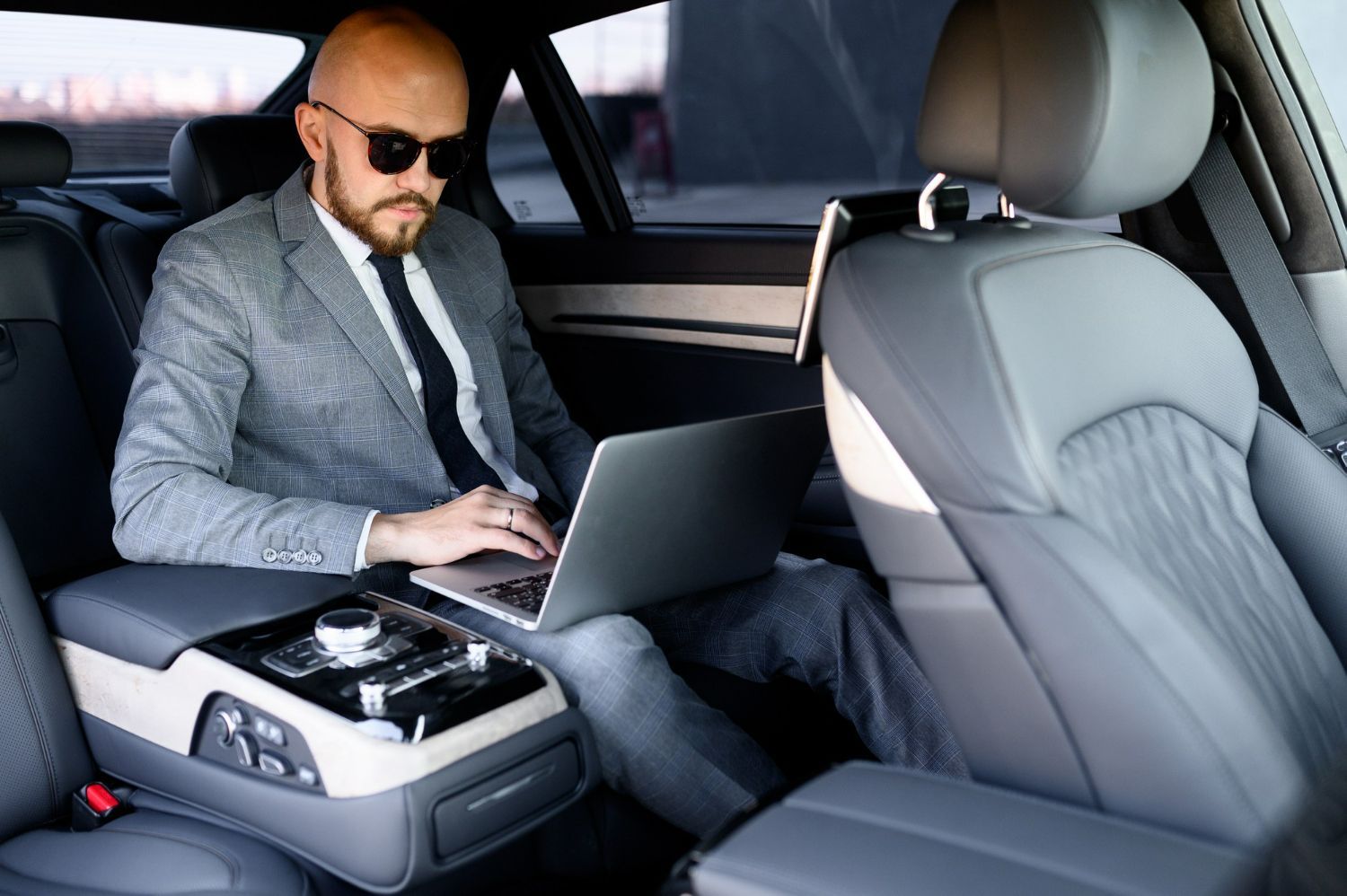 A man in a suit is sitting in the back seat of a car using a laptop computer.