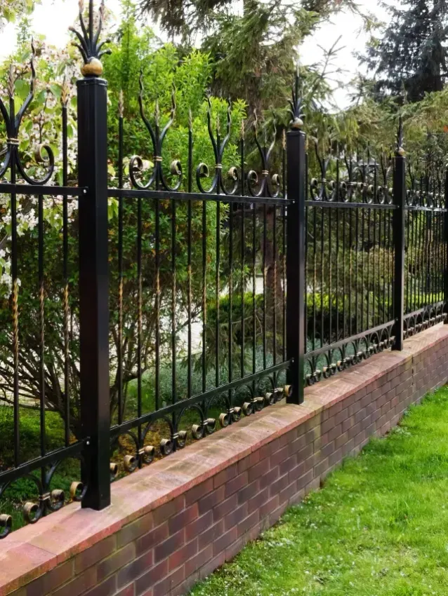 Fence Service El-Monte Ca - A decorative black iron fence with ornate details atop a brick wall, surrounded by lush greenery and colorful blooming plants.