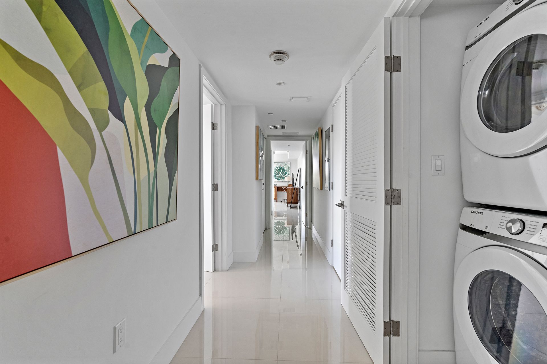 Narrow white hallway with colorful art, white doors, and stacked washer/dryer.