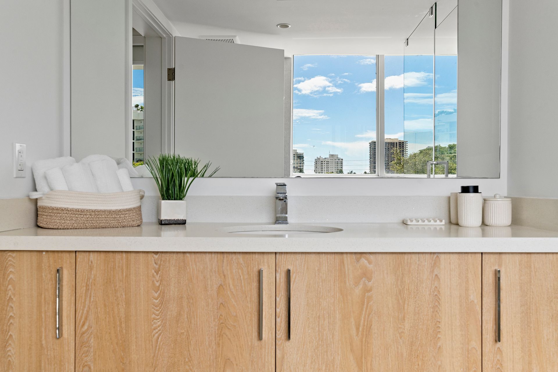 Bathroom with light wood cabinets, white countertop, large mirror, and view of buildings and sky.