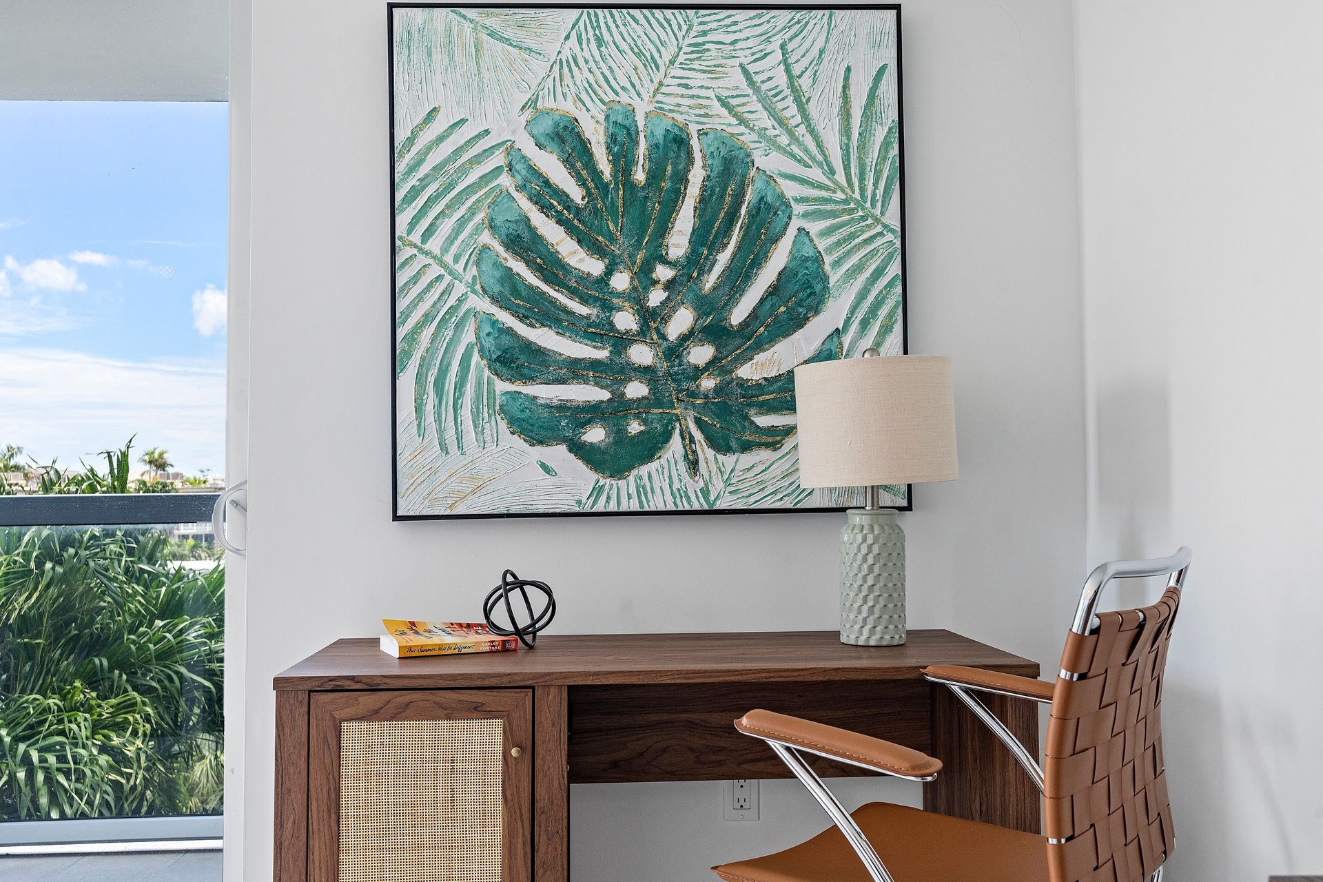 Desk with tropical leaf art, lamp, and woven leather chair in a bright room with a view.
