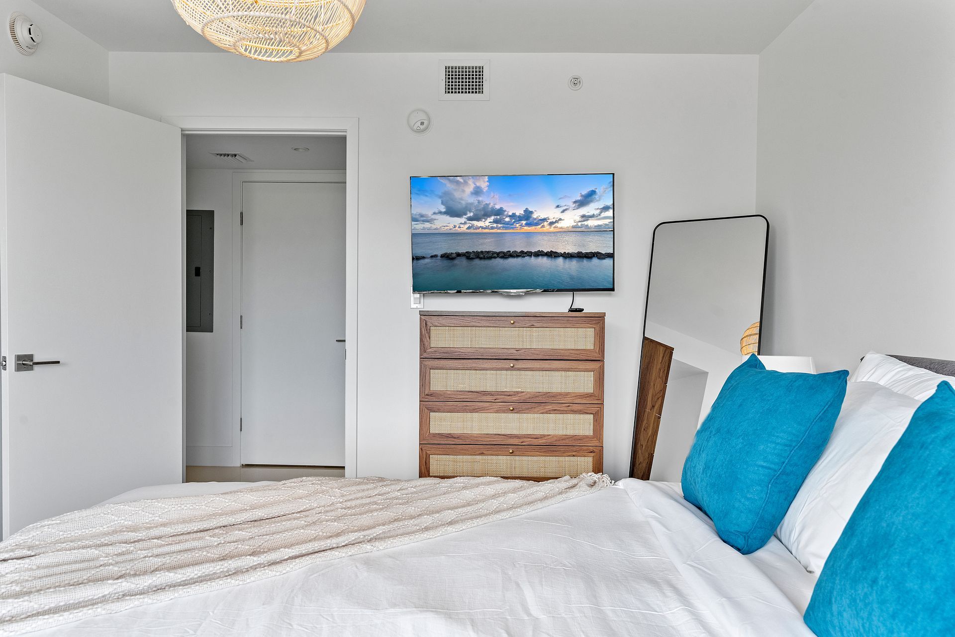 Bedroom with bed, dresser, TV, mirror, and open door to a hallway. White walls, teal pillows, and natural wood accents.